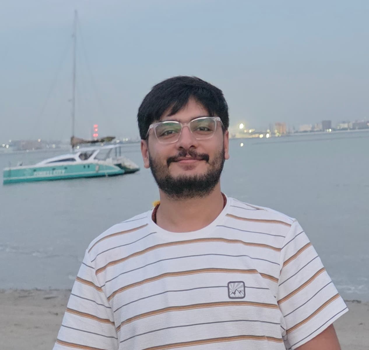 Takshak Mudgal smiling in clear-framed glasses and a striped T-shirt on a beach at dusk, with a catamaran on the water behind him